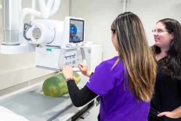 An FSW Radiologic Technology student in purple scrubs operates digital imaging equipment while a faculty member observes her.