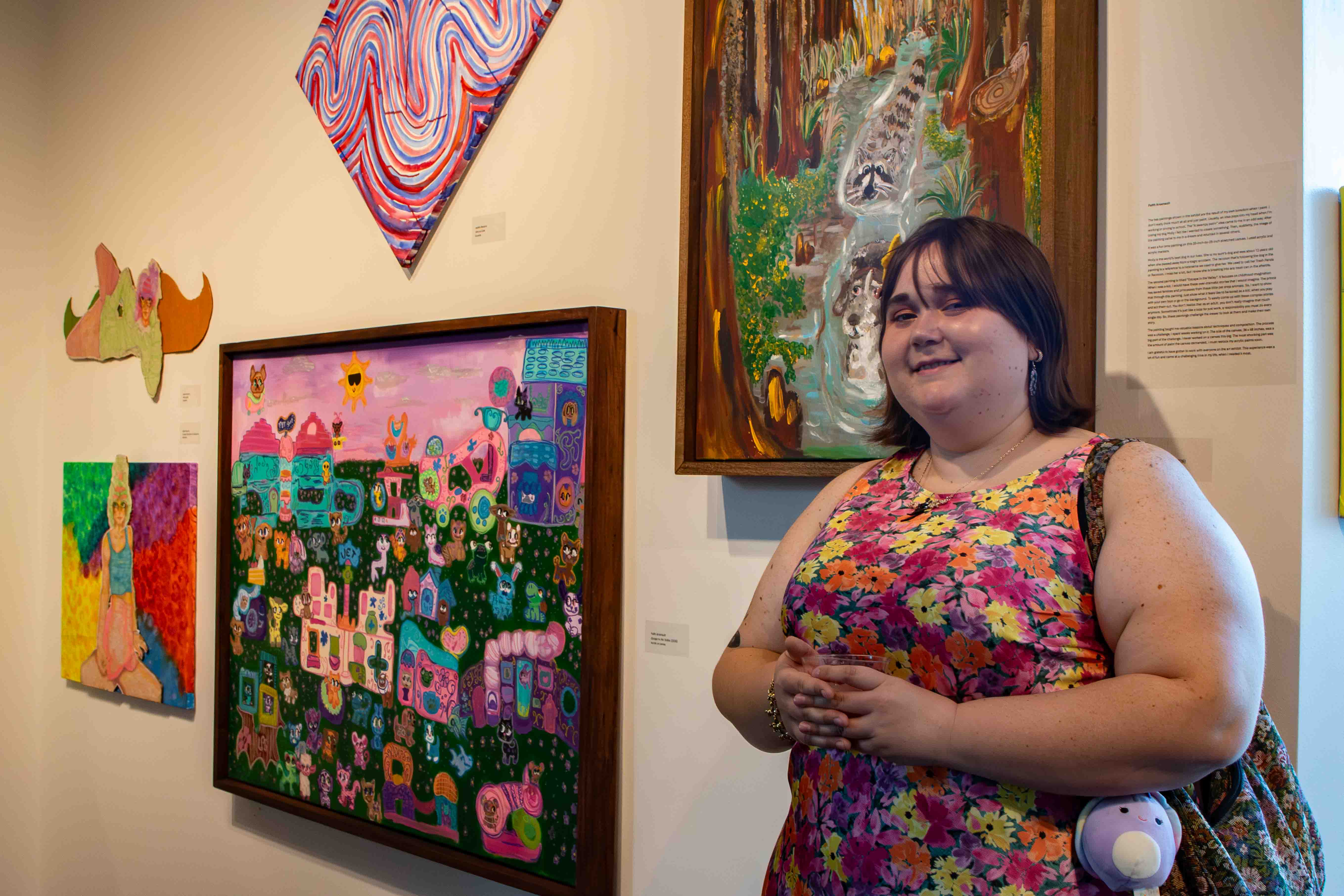 Faith Arsenault poses in front of her artwork and artist statement. Faith’s paintings drew heavily from her childhood toys and memories of being bored and using her imagination to entertain herself. Her paintings feature nearly 100 unique characters.