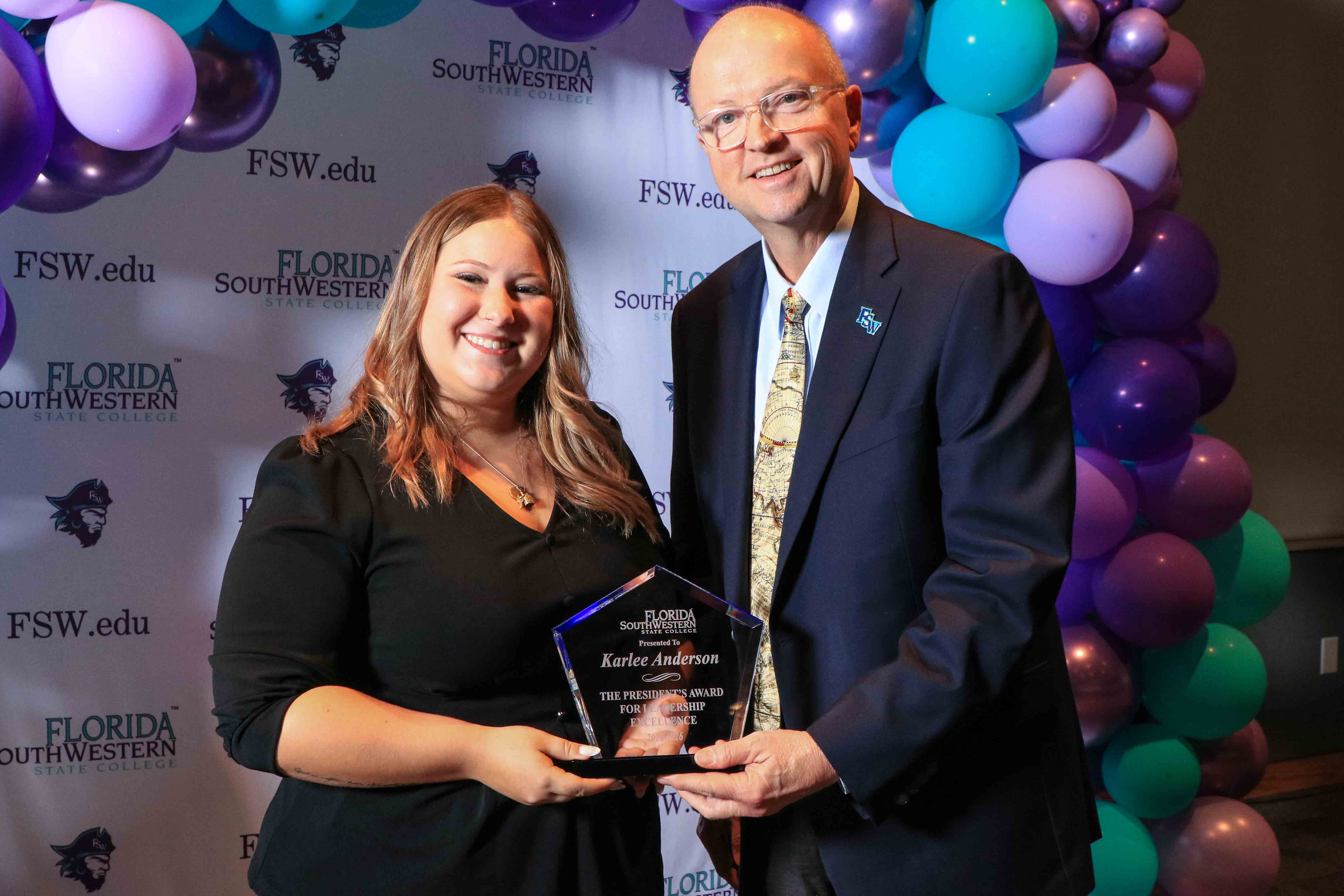 FSW President Dr. Jeff Allbritten presents the President's Award for Leadership Excellence to Karlee Anderson at the Lee Campus Life Awards on April 14, 2026 standing in front of a banner with FSW logos and a purple and aqua balloon arch.