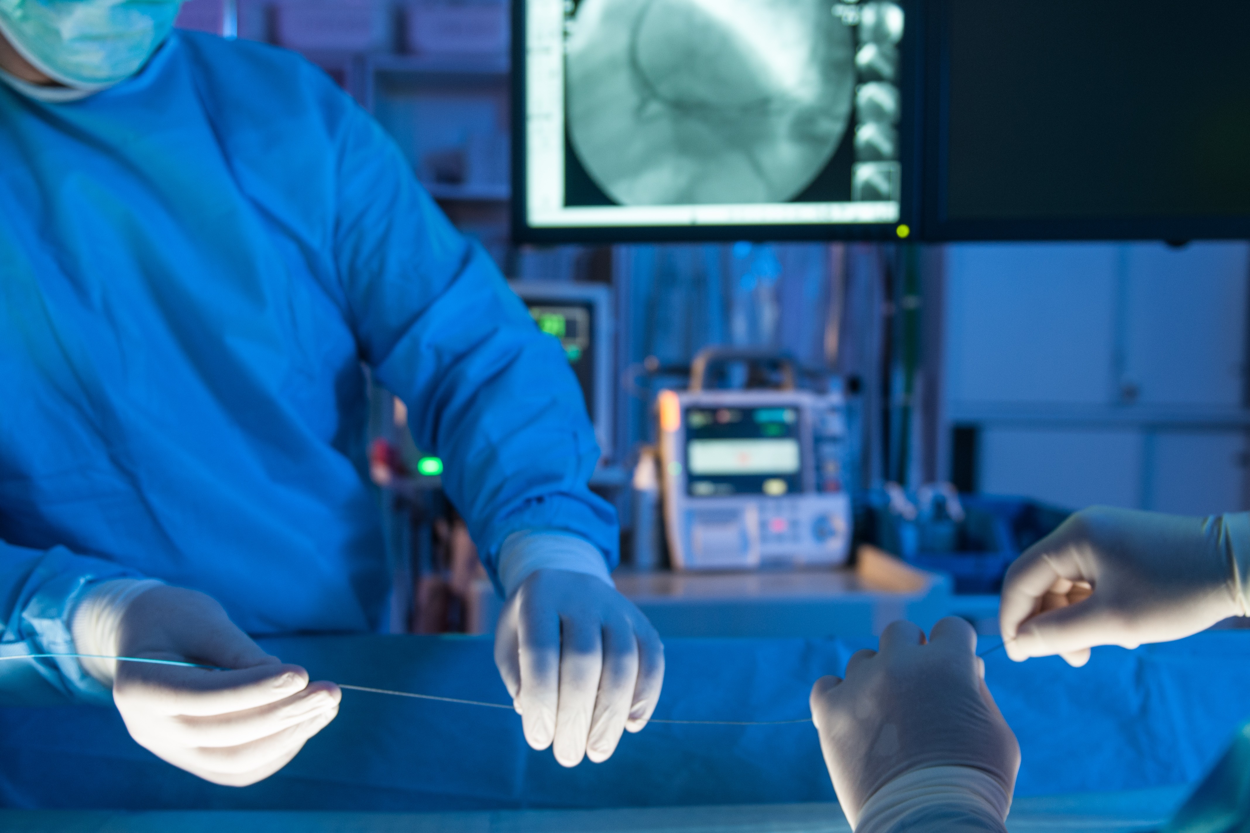 Close-up of gloved cardiovascular technologist performing a catheter-based procedure while medical imaging and monitoring screens are visible in the background