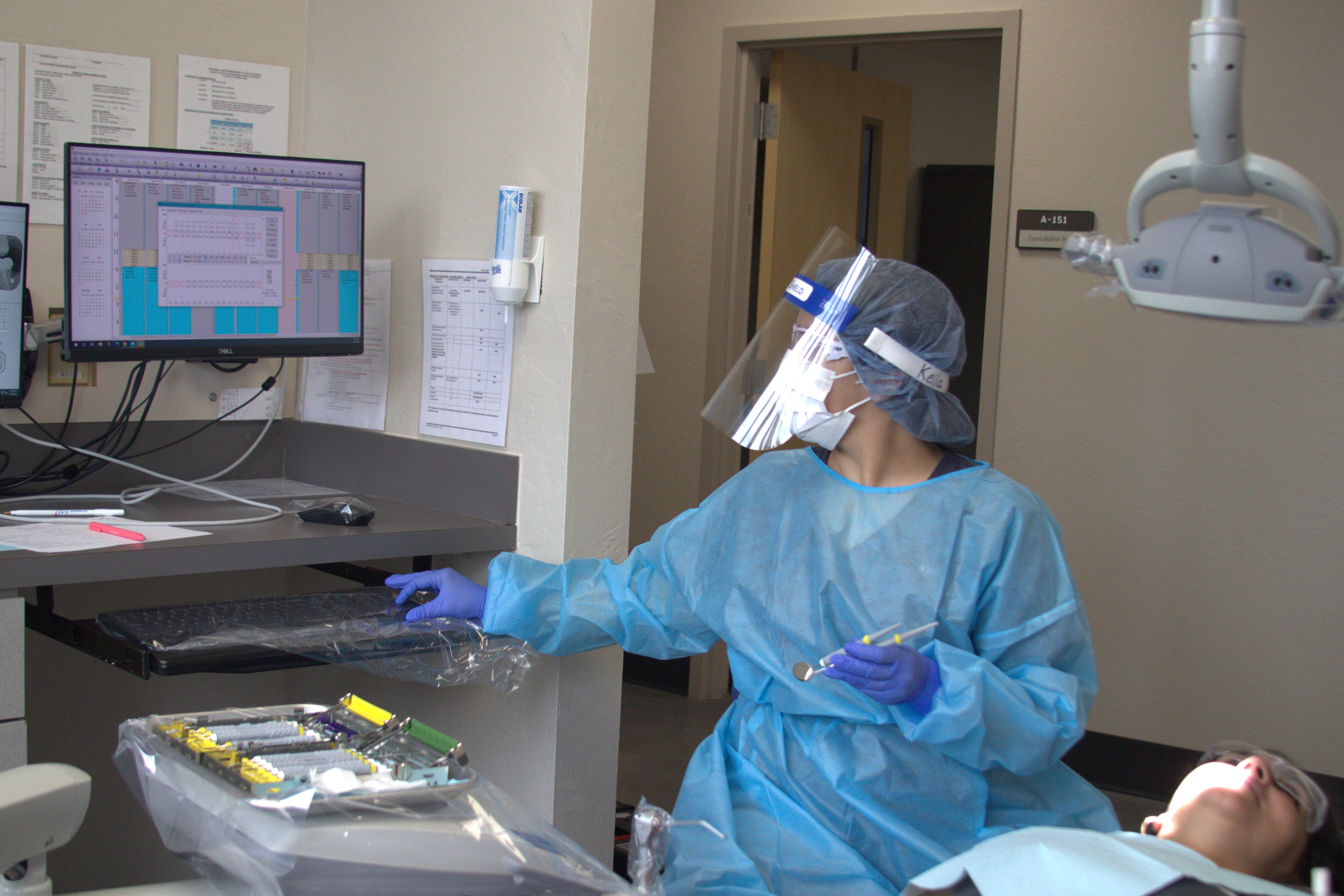 A dental hygiene student wearing a face shield, mask, gloves, and protective gown prepares dental instruments while looking at a computer monitor as a patient sits in a dental chair.