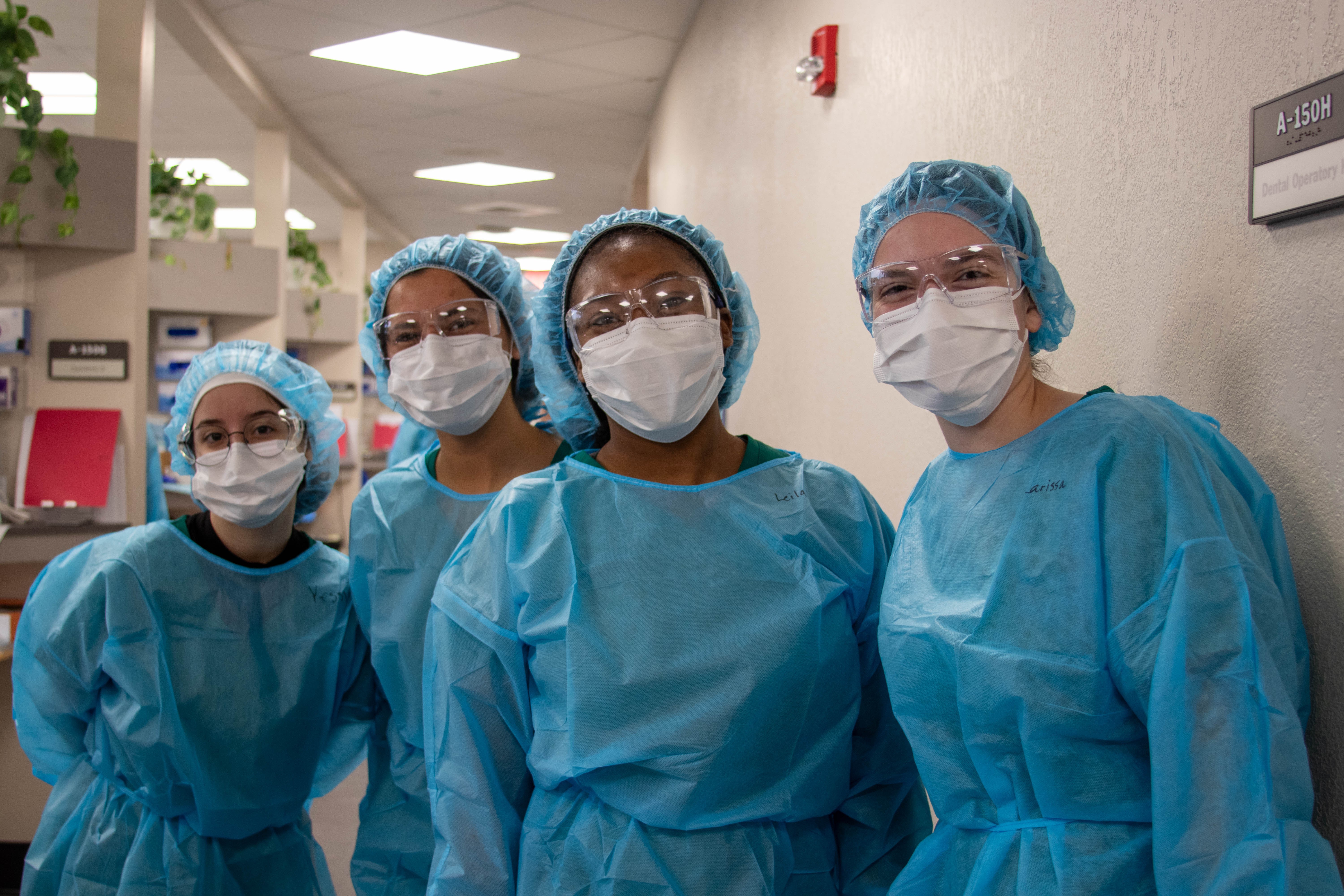 Four dental hygiene students wearing protective gowns, masks, safety glasses, stand together in a dental clinic hallway, facing the camera.