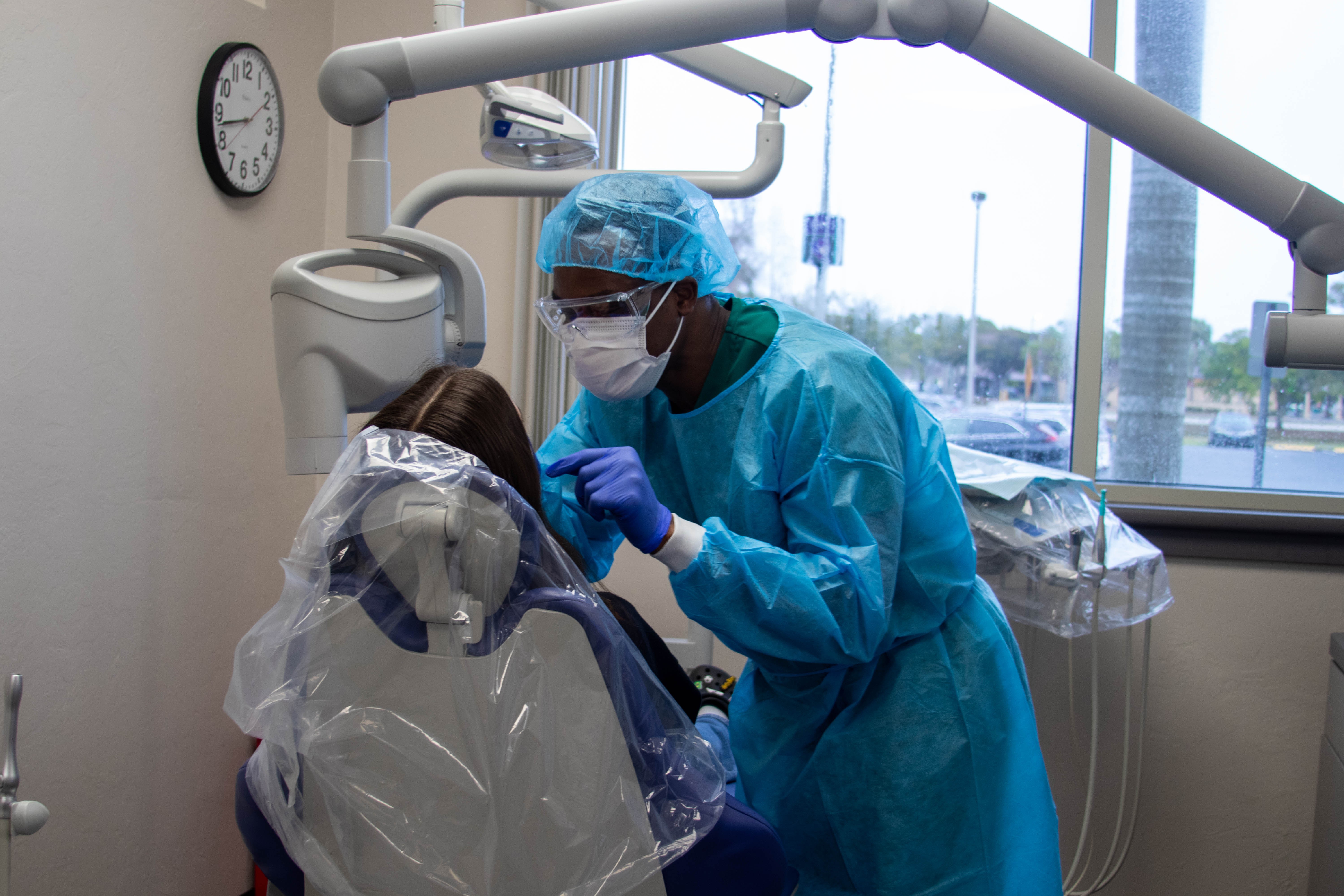 A dental hygiene student wearing a protective gown, mask, and safety glasses provides care to a patient seated in a dental chair.