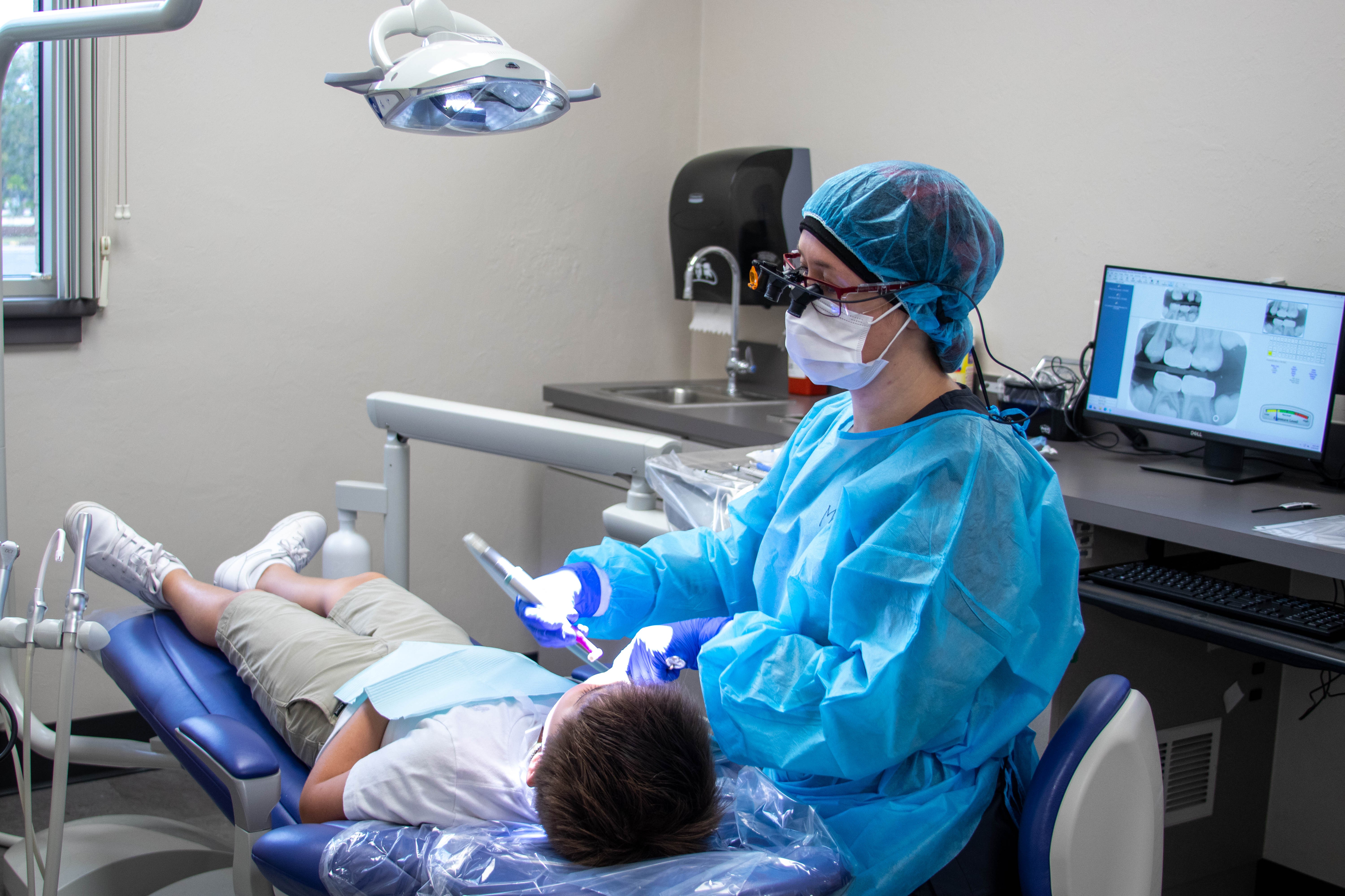 A dental student performs a cleaning on a patient reclined in a dental chair.