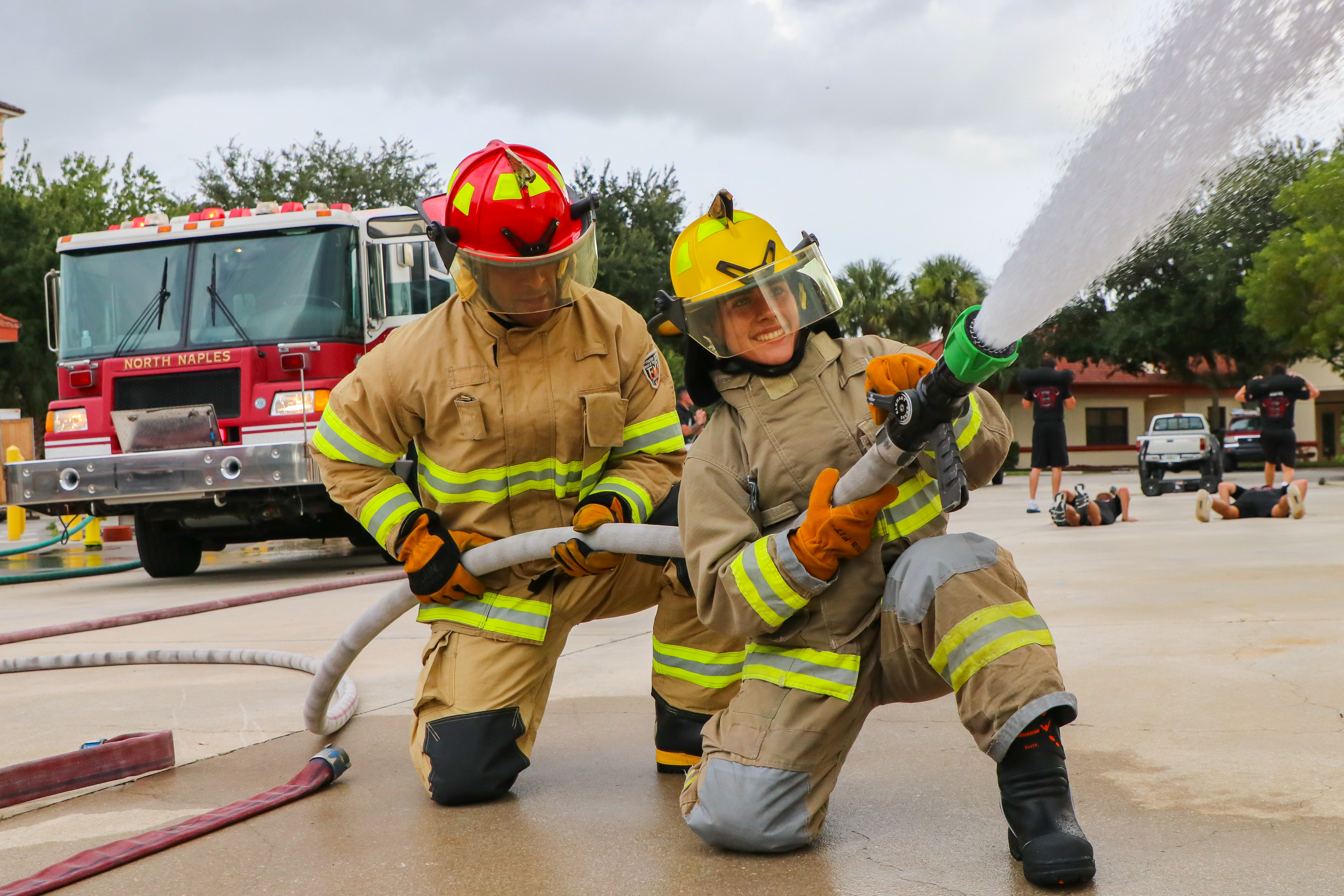 Fire instructor and a student in full gear kneel while spraying water from a fire hose during training.