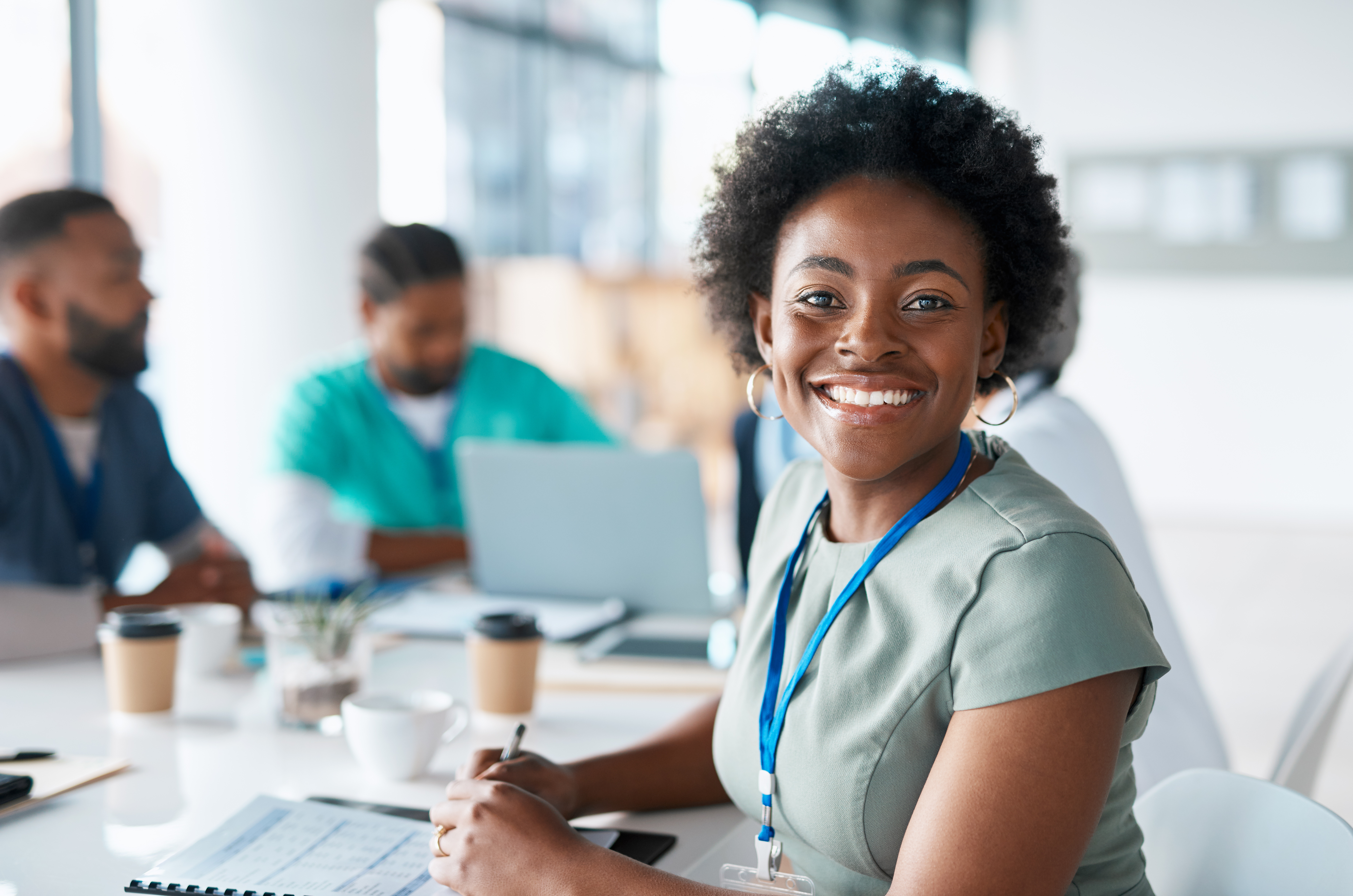 Woman smiling with a blue lanyard sits at a conference table holding a pen while coworkers work in the background.