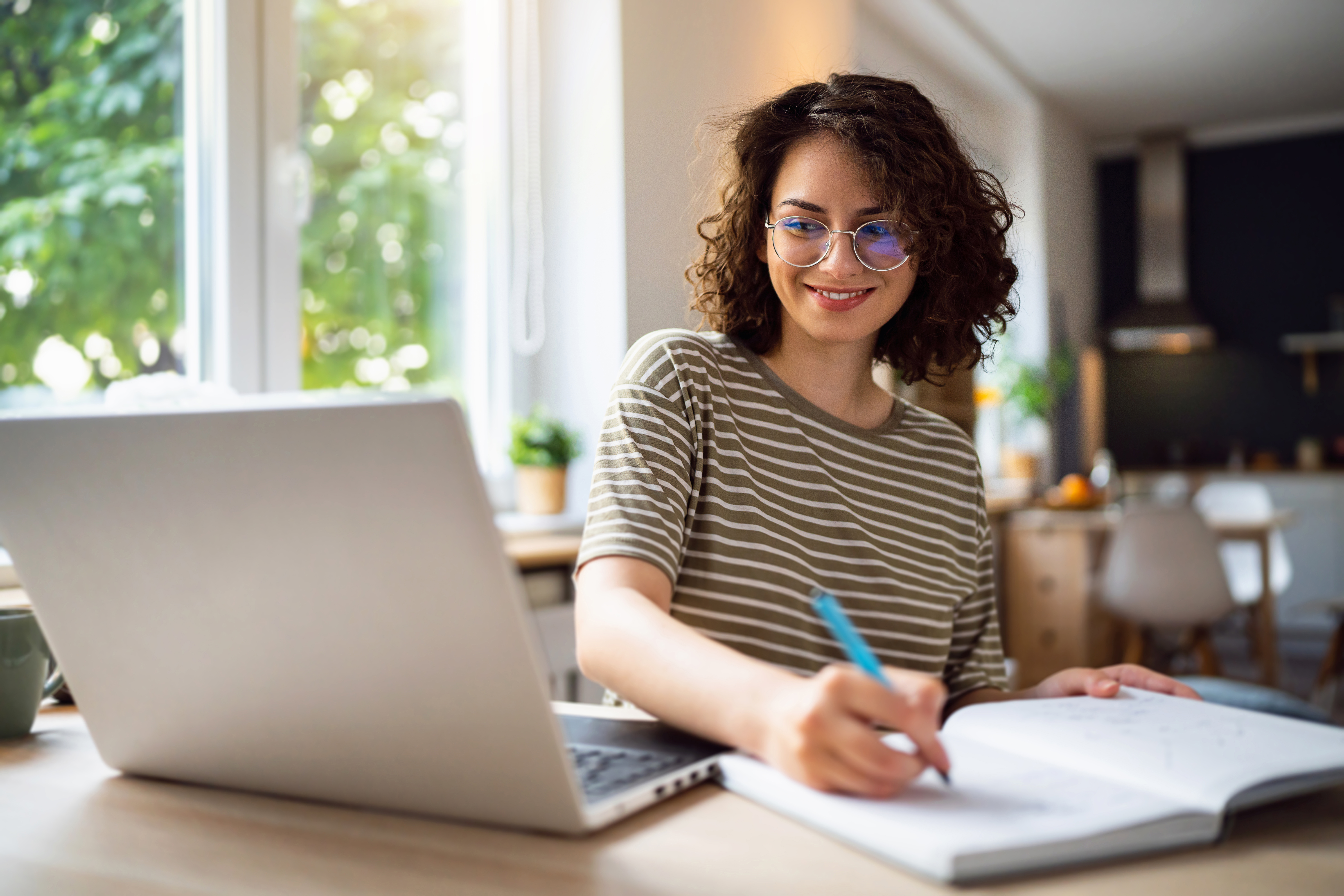 A young woman with curly hair and glasses smiles while looking at a laptop screen and taking notes in a notebook at a desk.