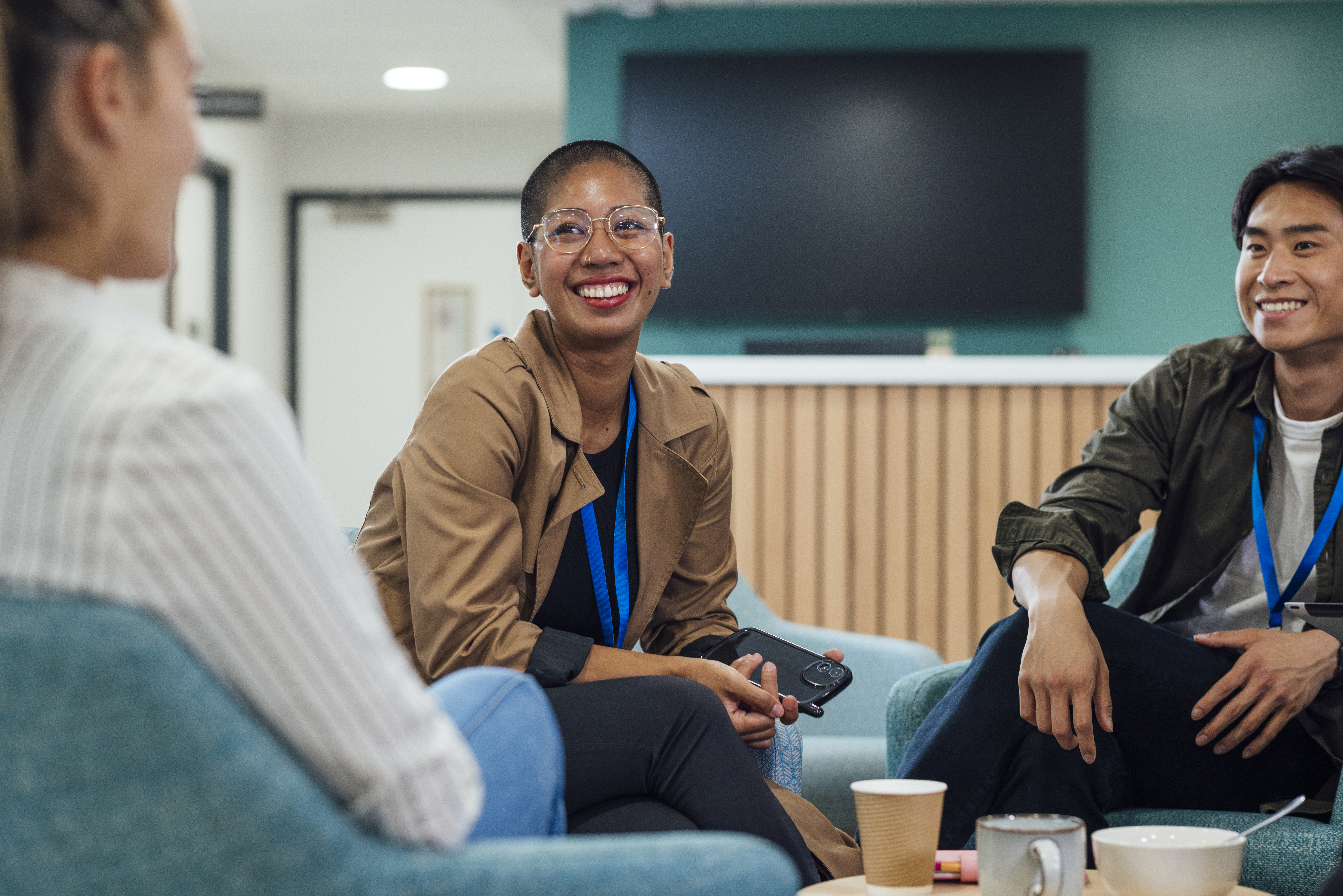Three coworkers wearing conference lanyards sit in a casual office lounge area smiling during a group conversation. A woman with glasses and a tan jacket sits in the center holding a phone while another coworker speaks in the foreground.