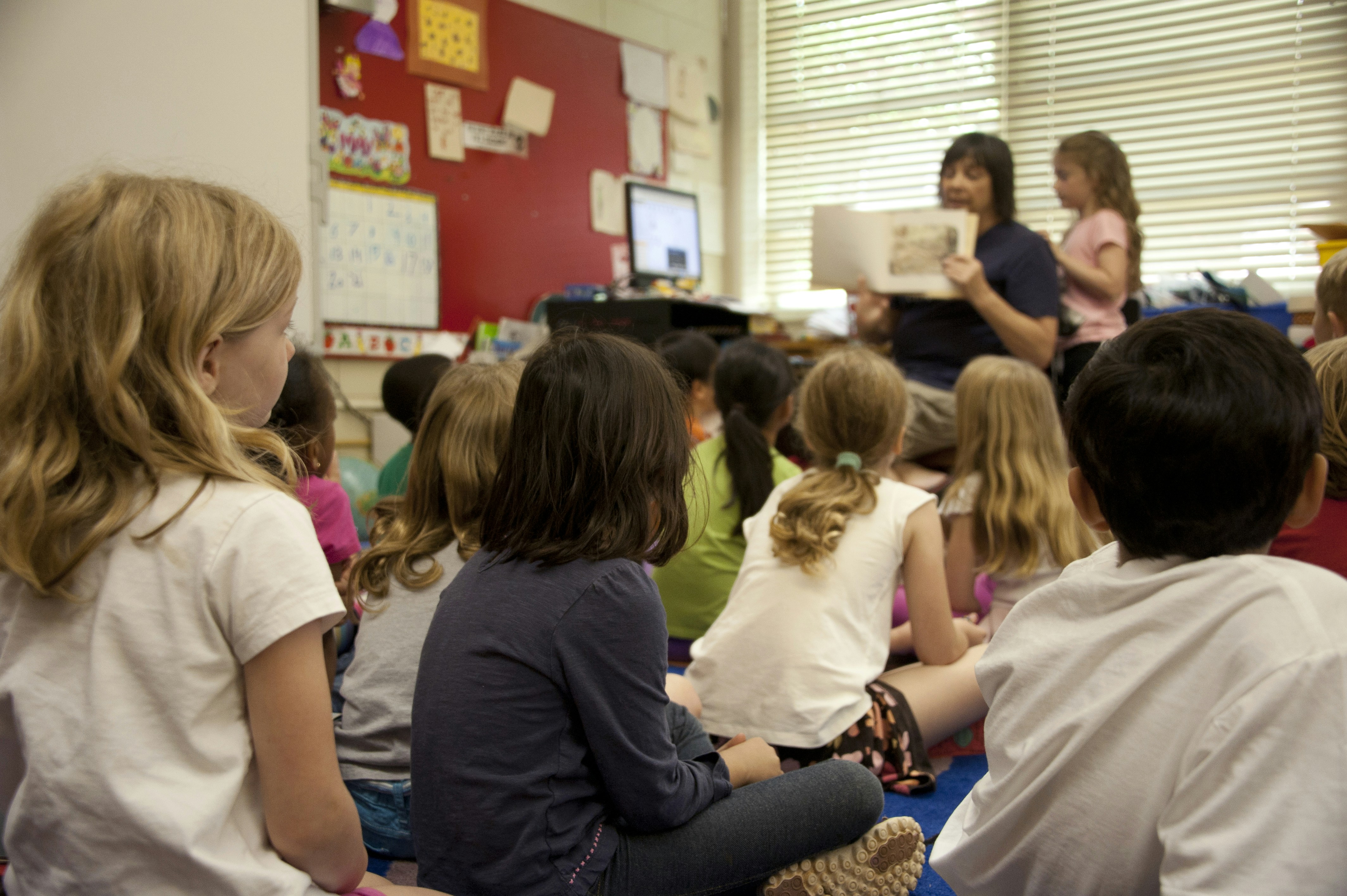 A group of young students sits cross-legged on the floor of a classroom, attentively listening to a teacher who is seated at the front holding up an illustrated storybook while reading aloud, with classroom decorations and educational materials on the walls.