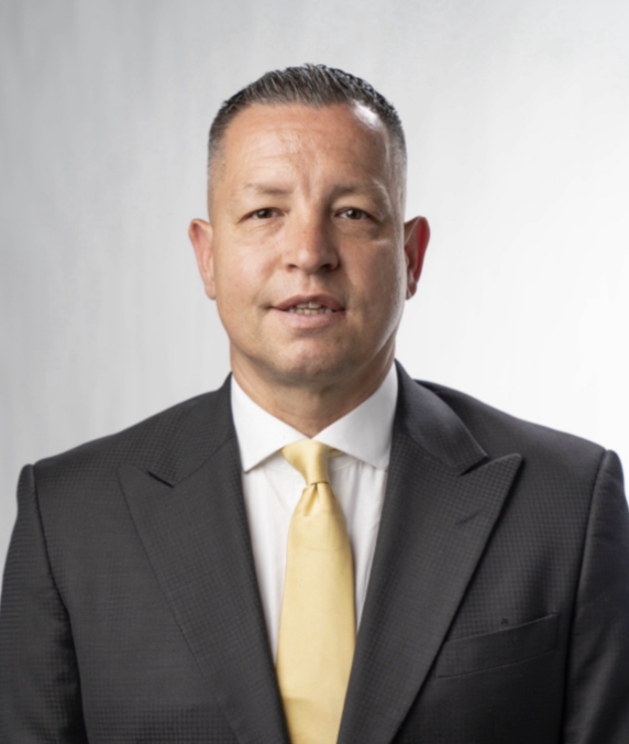 Headshot of David Grace, Head Coach of the Buccaneers Men's Basketball program, wearing a dark grey suit and yellow tie and smiling on a light gray background.