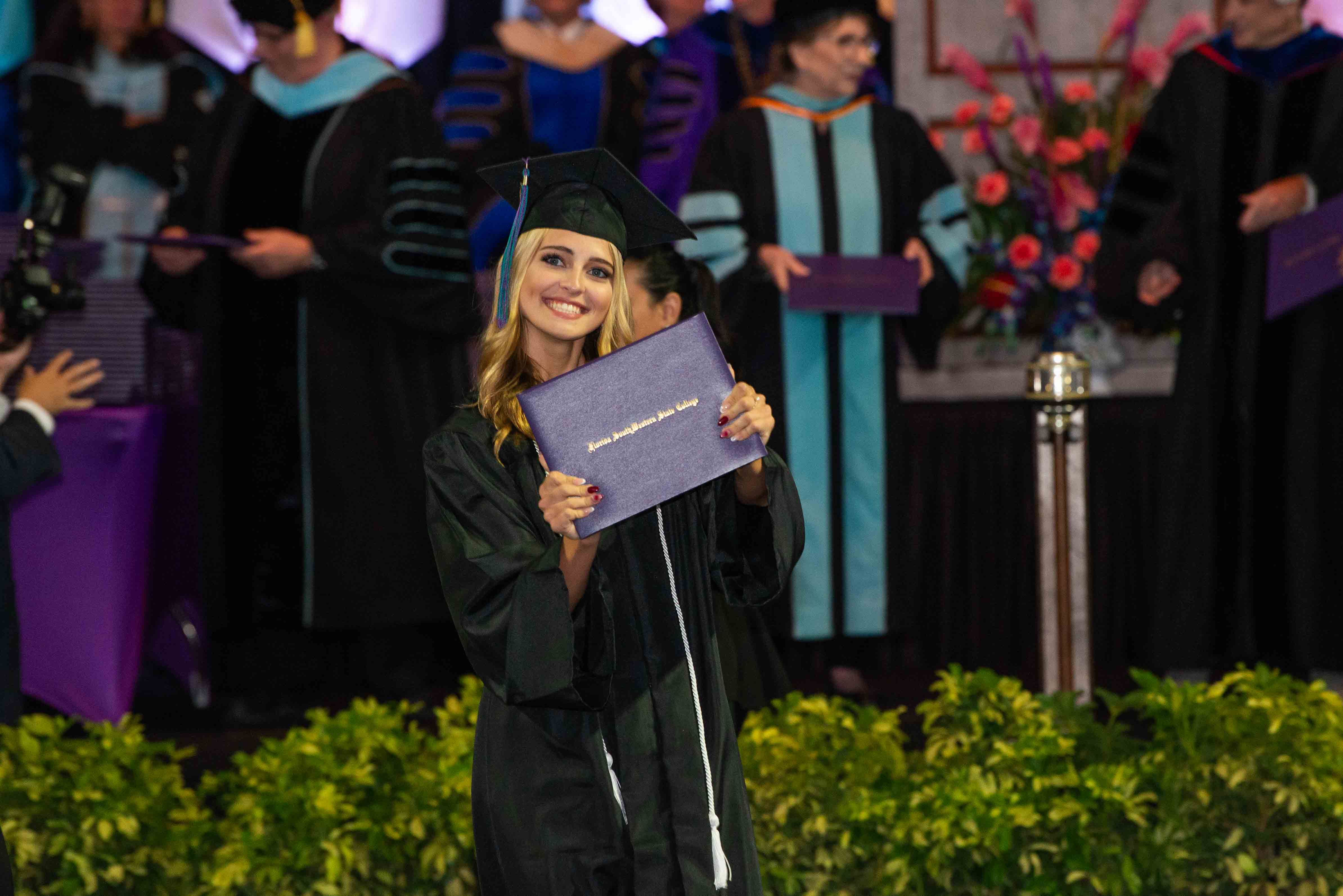 Smiling FSW graduate in cap and gown holding her Florida SouthWestern State College diploma cover at a commencement ceremony.