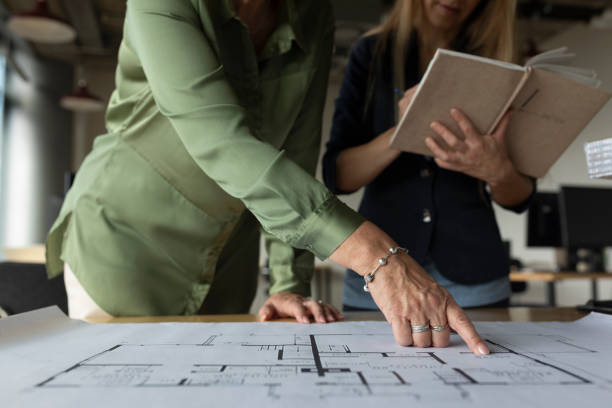 Two women reviewing architectural blueprints, with one pointing to a floor plan while the other takes notes.
