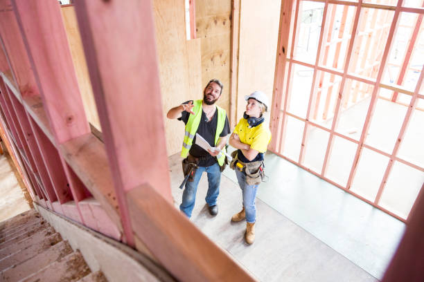 Two construction workers in safety gear reviewing plans and pointing upward inside a wooden-framed building.