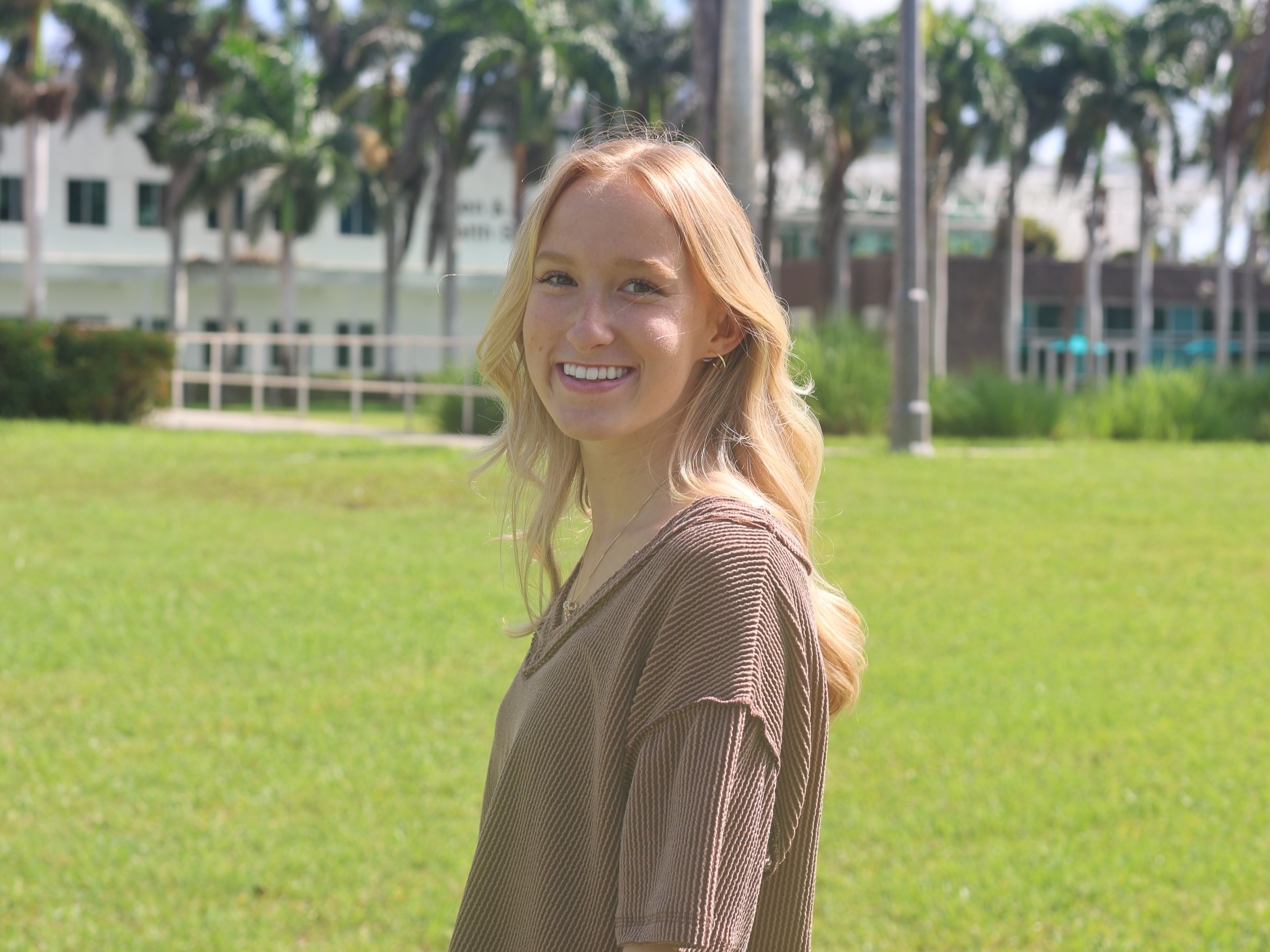 Shelby Williams, a student, poses smiling in front of the FSW Collier Campus entrance, with the campus building  in the background on a sunny day.